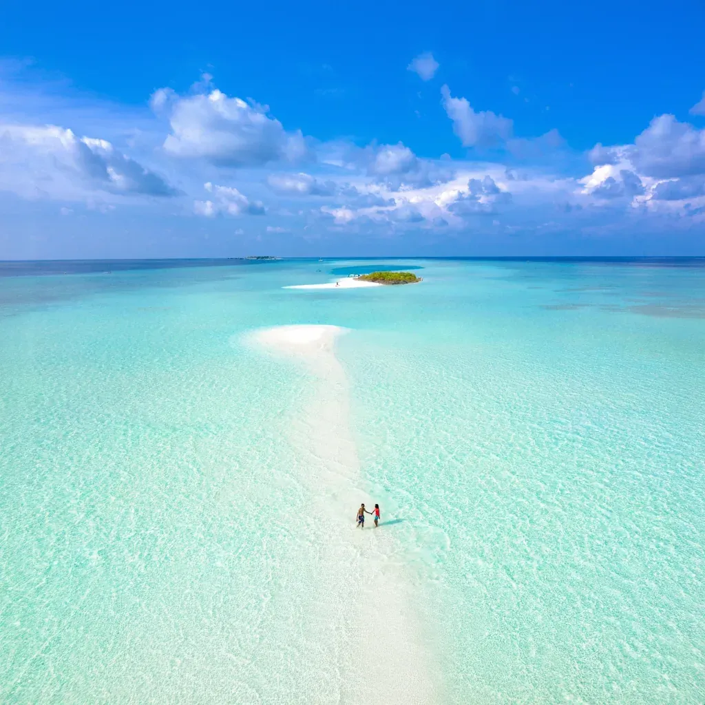 an aerial view of a long white sandbank curving through a shallow turquoise lagoon, two people walking along it, a small green island in the distance