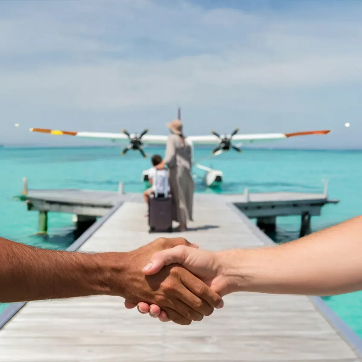 a close-up of two hands clasped in a handshake on a wooden jetty, a white and orange seaplane moored ahead with guests walking towards it with luggage, turquoise lagoon behind
