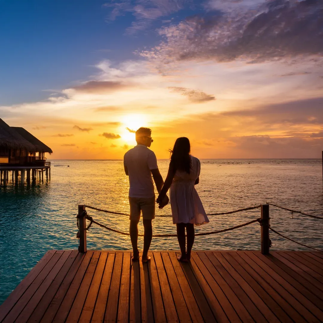 a couple from behind, holding hands at the end of a wooden jetty, the sky orange as the sun sets over the lagoon