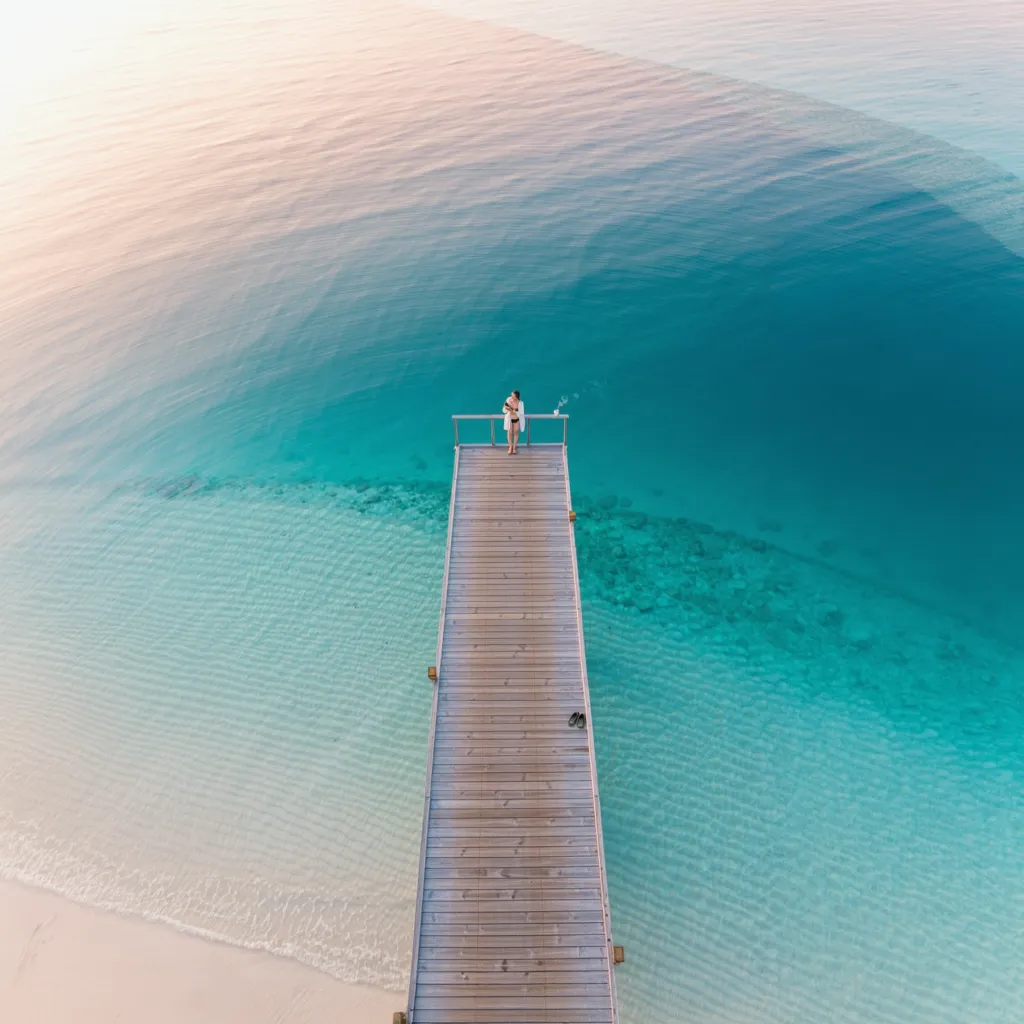 aerial view of a figure standing at the far end of a long wooden jetty reaching into a clear turquoise lagoon, sandals left on the boards behind them