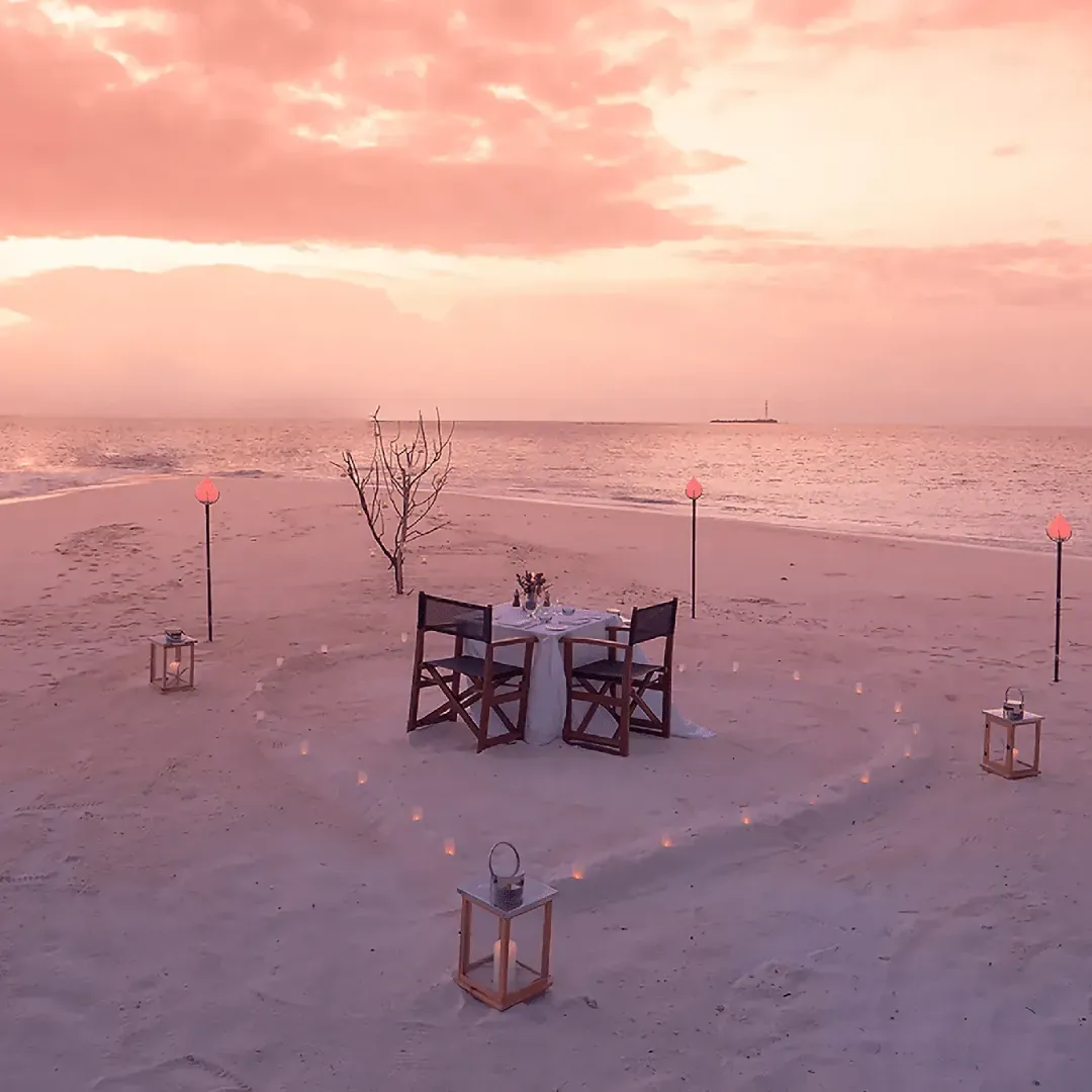 A private dinner table for two set on a sandbank at sunset, surrounded by lanterns and torches, with the ocean under a pink sky