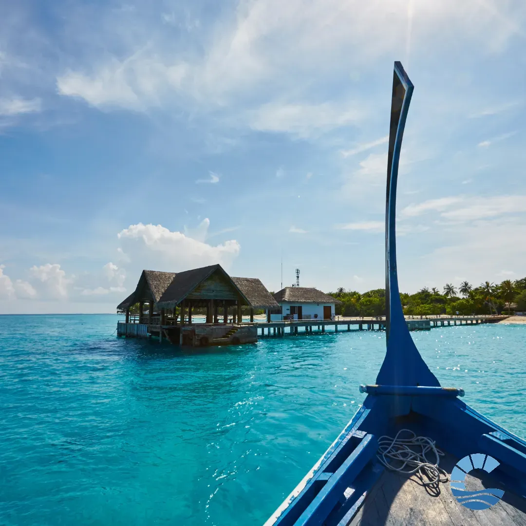The bow of a traditional blue dhoni boat approaching a local island jetty with a thatched-roof pavilion, turquoise lagoon and palm trees under a bright sky