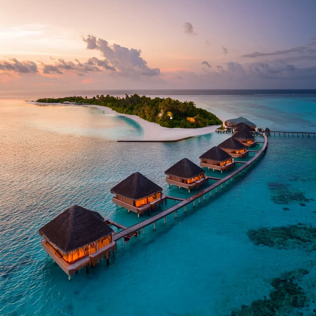 Aerial view of thatched overwater villas along a curved jetty at sunset, glowing warmly beside a small island with a white sand beach and turquoise lagoon